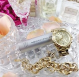A collection of luxury items including a gold watch, a chunky gold bracelet, and a silver canister of Rodan + Fields Active Hydration Serum. These items are placed on a crystal dish surrounded by pink rose petals and several perfume bottles, creating an elegant and sophisticated arrangement.