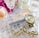 A collection of luxury items including a gold watch, a chunky gold bracelet, and a silver canister of Rodan + Fields Active Hydration Serum. These items are placed on a crystal dish surrounded by pink rose petals and several perfume bottles, creating an elegant and sophisticated arrangement.