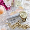 A collection of luxury items including a gold watch, a chunky gold bracelet, and a silver canister of Rodan + Fields Active Hydration Serum. These items are placed on a crystal dish surrounded by pink rose petals and several perfume bottles, creating an elegant and sophisticated arrangement.