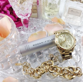 A collection of luxury items including a gold watch, a chunky gold bracelet, and a silver canister of Rodan + Fields Active Hydration Serum. These items are placed on a crystal dish surrounded by pink rose petals and several perfume bottles, creating an elegant and sophisticated arrangement.