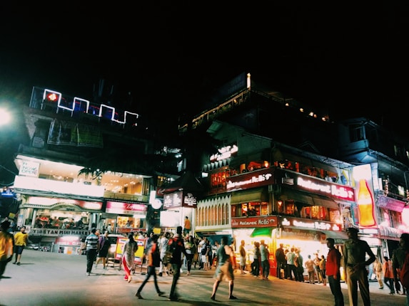 A bustling nightlife scene with a lively crowd on a brightly lit street. Various shops and restaurants with illuminated signs create a vibrant atmosphere. People are casually strolling or gathered around, enhancing the dynamic and energetic setting. The street is busy with activity, featuring a mix of locals and tourists.