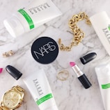 A flat lay of beauty and fashion items on a marble surface, including a tube of Rodan + Fields Soothe gentle cream wash, a container of NARS product, a MAC lipstick with the cap off showing a pink shade, a gold chain bracelet, a gold wristwatch, a pair of small earrings, and two thin gold rings.