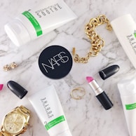 A flat lay of beauty and fashion items on a marble surface, including a tube of Rodan + Fields Soothe gentle cream wash, a container of NARS product, a MAC lipstick with the cap off showing a pink shade, a gold chain bracelet, a gold wristwatch, a pair of small earrings, and two thin gold rings.