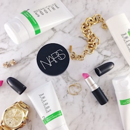 A flat lay of beauty and fashion items on a marble surface, including a tube of Rodan + Fields Soothe gentle cream wash, a container of NARS product, a MAC lipstick with the cap off showing a pink shade, a gold chain bracelet, a gold wristwatch, a pair of small earrings, and two thin gold rings.