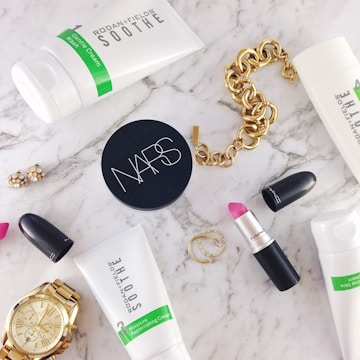 A flat lay of beauty and fashion items on a marble surface, including a tube of Rodan + Fields Soothe gentle cream wash, a container of NARS product, a MAC lipstick with the cap off showing a pink shade, a gold chain bracelet, a gold wristwatch, a pair of small earrings, and two thin gold rings.