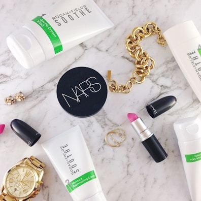 A flat lay of beauty and fashion items on a marble surface, including a tube of Rodan + Fields Soothe gentle cream wash, a container of NARS product, a MAC lipstick with the cap off showing a pink shade, a gold chain bracelet, a gold wristwatch, a pair of small earrings, and two thin gold rings.