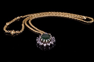 A delicate gold necklace with a prominent emerald green gemstone pendant surrounded by small clear crystals. The necklace is elegantly arranged on a black background, highlighting the lustrous gold chain and the vibrant green jewel.