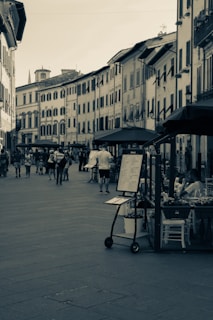 A narrow European street is lined with old-style buildings featuring shutters on the windows. People are walking along the street, some appearing to browse the menus and items displayed outside a restaurant with outdoor seating. The atmosphere suggests a leisurely pace typical of a picturesque European town.