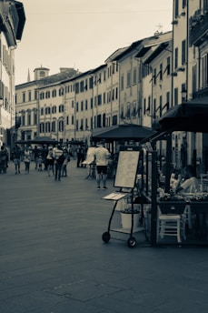 A narrow European street is lined with old-style buildings featuring shutters on the windows. People are walking along the street, some appearing to browse the menus and items displayed outside a restaurant with outdoor seating. The atmosphere suggests a leisurely pace typical of a picturesque European town.