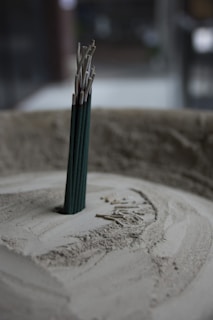 A group of incense sticks standing upright in a circular container filled with sand. The incense sticks are partially burned, with ash collected around the base. The soft lighting and muted colors create a tranquil atmosphere.