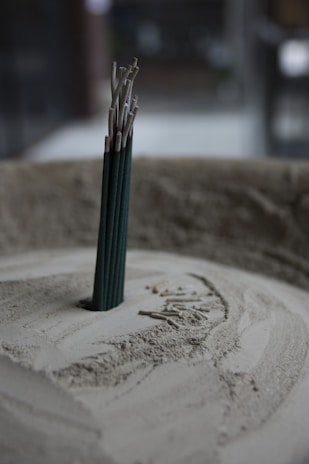 A group of incense sticks standing upright in a circular container filled with sand. The incense sticks are partially burned, with ash collected around the base. The soft lighting and muted colors create a tranquil atmosphere.