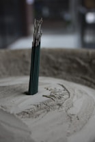 A group of incense sticks standing upright in a circular container filled with sand. The incense sticks are partially burned, with ash collected around the base. The soft lighting and muted colors create a tranquil atmosphere.