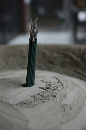 A group of incense sticks standing upright in a circular container filled with sand. The incense sticks are partially burned, with ash collected around the base. The soft lighting and muted colors create a tranquil atmosphere.