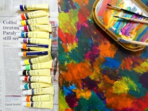 A collection of acrylic paint tubes is arranged on a newspaper. Beside them lies a colorful palette covered with various shades of paint, and several paintbrushes rest on top. The colors are vibrant and diverse, with an abstract background of mixed hues.