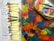 A collection of acrylic paint tubes is arranged on a newspaper. Beside them lies a colorful palette covered with various shades of paint, and several paintbrushes rest on top. The colors are vibrant and diverse, with an abstract background of mixed hues.