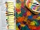 A collection of acrylic paint tubes is arranged on a newspaper. Beside them lies a colorful palette covered with various shades of paint, and several paintbrushes rest on top. The colors are vibrant and diverse, with an abstract background of mixed hues.