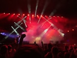 A vibrant concert scene with a crowd of people enjoying a live performance. Bright red and white stage lights illuminate the stage, creating a dynamic atmosphere. Smoke effects enhance the dramatic ambiance as the audience raises their hands, showing enthusiasm and engagement.