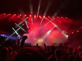 A vibrant concert scene with a crowd of people enjoying a live performance. Bright red and white stage lights illuminate the stage, creating a dynamic atmosphere. Smoke effects enhance the dramatic ambiance as the audience raises their hands, showing enthusiasm and engagement.