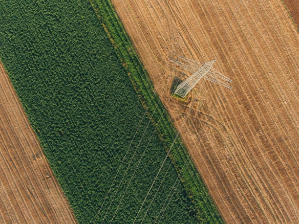 An aerial view of agricultural fields divided by a line of green crops on one side and brown, recently plowed soil on the other. A power transmission tower stands prominently at the intersection of these two segments, with power lines extending across the fields.