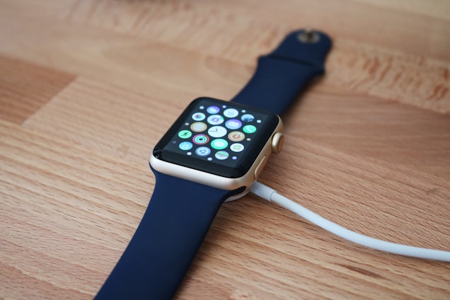 A smartwatch with a gold-colored frame and a dark blue band is placed on a wooden surface. The screen displays multiple colorful app icons, and a white charging cable is connected to it.