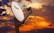 A large white satellite dish labeled 'TV SAT' is prominently positioned against a dramatic sunset sky filled with vibrant orange, purple, and blue hues. The satellite dish is mounted on a sturdy stand and angled upward, capturing signals.