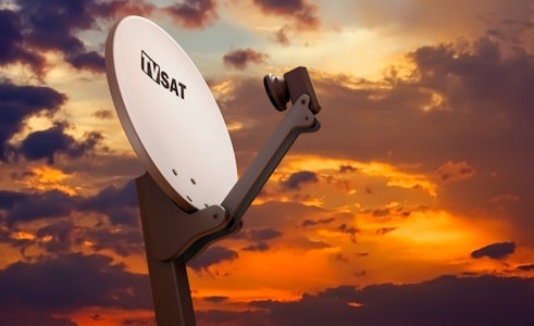 A large white satellite dish labeled 'TV SAT' is prominently positioned against a dramatic sunset sky filled with vibrant orange, purple, and blue hues. The satellite dish is mounted on a sturdy stand and angled upward, capturing signals.