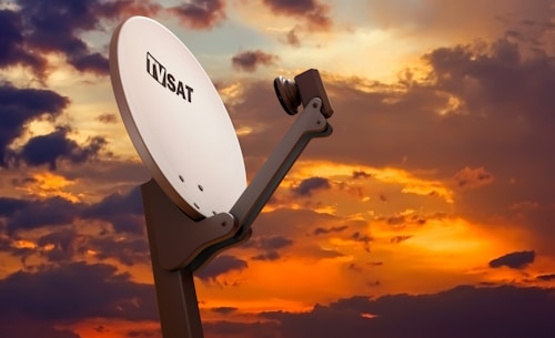 A large white satellite dish labeled 'TV SAT' is prominently positioned against a dramatic sunset sky filled with vibrant orange, purple, and blue hues. The satellite dish is mounted on a sturdy stand and angled upward, capturing signals.