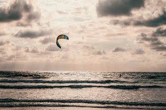 A kite surfer rides the ocean waves under a cloudy sky. The kite is multicolored and stands out against the overcast background. The sun reflects off the water, creating a sparkling effect.