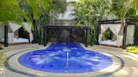A luxurious outdoor setting featuring a circular blue jacuzzi or small pool with gentle bubbles, surrounded by a stone pathway. Two cabanas with white draped curtains and lounge beds on either side of the pool create a sense of relaxation and privacy. The area is lush with surrounding palm trees and tropical plants, adding to the tranquil atmosphere.