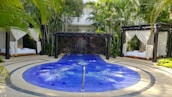A luxurious outdoor setting featuring a circular blue jacuzzi or small pool with gentle bubbles, surrounded by a stone pathway. Two cabanas with white draped curtains and lounge beds on either side of the pool create a sense of relaxation and privacy. The area is lush with surrounding palm trees and tropical plants, adding to the tranquil atmosphere.