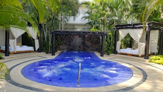 A luxurious outdoor setting featuring a circular blue jacuzzi or small pool with gentle bubbles, surrounded by a stone pathway. Two cabanas with white draped curtains and lounge beds on either side of the pool create a sense of relaxation and privacy. The area is lush with surrounding palm trees and tropical plants, adding to the tranquil atmosphere.
