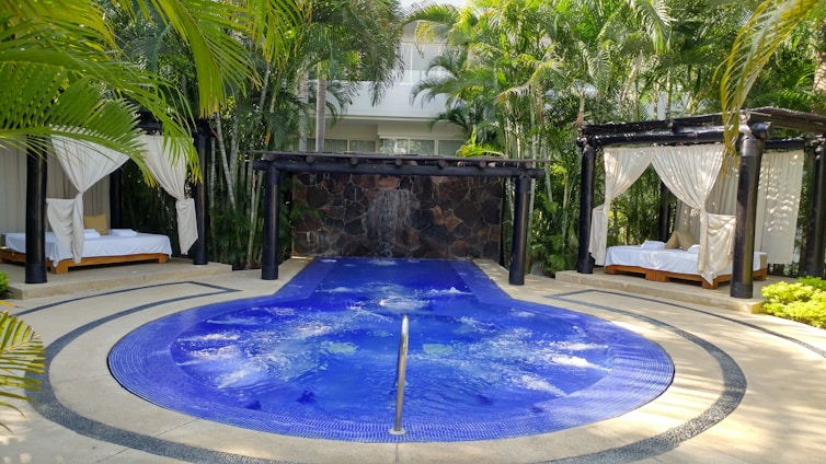 A luxurious outdoor setting featuring a circular blue jacuzzi or small pool with gentle bubbles, surrounded by a stone pathway. Two cabanas with white draped curtains and lounge beds on either side of the pool create a sense of relaxation and privacy. The area is lush with surrounding palm trees and tropical plants, adding to the tranquil atmosphere.