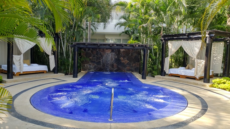A luxurious outdoor setting featuring a circular blue jacuzzi or small pool with gentle bubbles, surrounded by a stone pathway. Two cabanas with white draped curtains and lounge beds on either side of the pool create a sense of relaxation and privacy. The area is lush with surrounding palm trees and tropical plants, adding to the tranquil atmosphere.