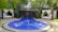 A luxurious outdoor setting featuring a circular blue jacuzzi or small pool with gentle bubbles, surrounded by a stone pathway. Two cabanas with white draped curtains and lounge beds on either side of the pool create a sense of relaxation and privacy. The area is lush with surrounding palm trees and tropical plants, adding to the tranquil atmosphere.