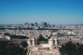 A panoramic view of a large cityscape with a mix of historical and modern architecture. In the foreground, there's a notable structure with columns surrounded by lush green trees, likely a public square or park area. The background showcases a skyline filled with modern skyscrapers under a clear blue sky.