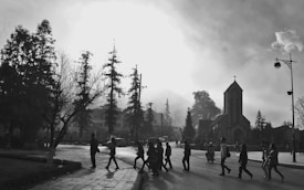 A black and white scene of several people walking across a street in front of a church with a prominent steeple. Tall trees and street lamps line the road, and the atmosphere is misty, creating a dramatic effect with the bright sky.