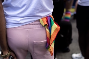 A person wearing a white shirt and pink pants stands in a crowd. A colorful rainbow flag is tucked into the back pocket of the pants. The setting suggests an outdoor event with people dressed in casual clothing. There is a sense of diversity and inclusivity.