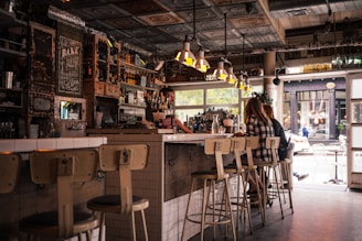 A cozy, rustic café interior featuring wooden and metal furnishings with a bar counter lined with stools. The space is decorated with vintage elements, such as a chalkboard menu and various bottles on shelves. Customers sit at the bar, engaging in conversation, with an inviting atmosphere enhanced by warm lighting from overhead lamps. The large window allows natural light to filter in, offering a view of the street outside.