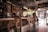 A cozy, rustic café interior featuring wooden and metal furnishings with a bar counter lined with stools. The space is decorated with vintage elements, such as a chalkboard menu and various bottles on shelves. Customers sit at the bar, engaging in conversation, with an inviting atmosphere enhanced by warm lighting from overhead lamps. The large window allows natural light to filter in, offering a view of the street outside.