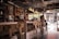 A cozy, rustic café interior featuring wooden and metal furnishings with a bar counter lined with stools. The space is decorated with vintage elements, such as a chalkboard menu and various bottles on shelves. Customers sit at the bar, engaging in conversation, with an inviting atmosphere enhanced by warm lighting from overhead lamps. The large window allows natural light to filter in, offering a view of the street outside.