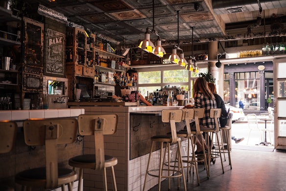 A cozy, rustic café interior featuring wooden and metal furnishings with a bar counter lined with stools. The space is decorated with vintage elements, such as a chalkboard menu and various bottles on shelves. Customers sit at the bar, engaging in conversation, with an inviting atmosphere enhanced by warm lighting from overhead lamps. The large window allows natural light to filter in, offering a view of the street outside.