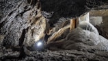 A rugged cave interior with textured rock formations and stalactites. There is a spotlight illuminating the surface, highlighting the rough, uneven textures and the damp, shiny surfaces of the cave walls and formations.