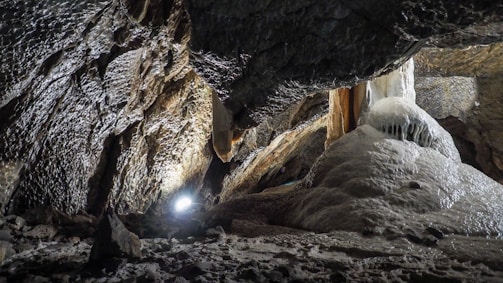 A rugged cave interior with textured rock formations and stalactites. There is a spotlight illuminating the surface, highlighting the rough, uneven textures and the damp, shiny surfaces of the cave walls and formations.