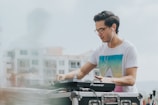 A person wearing a white t-shirt with a colorful geometric design is using DJ equipment, likely mixing music. They have headphones on and seem focused on their task. The background shows a blurred view of buildings and a microphone is present in the foreground.