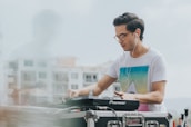A person wearing a white t-shirt with a colorful geometric design is using DJ equipment, likely mixing music. They have headphones on and seem focused on their task. The background shows a blurred view of buildings and a microphone is present in the foreground.