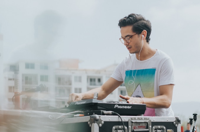 A person wearing a white t-shirt with a colorful geometric design is using DJ equipment, likely mixing music. They have headphones on and seem focused on their task. The background shows a blurred view of buildings and a microphone is present in the foreground.