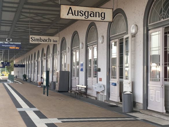 A train station platform with arched doorways and large windows along the right side. Signs hanging from the ceiling indicate 'Ausgang' (exit) and 'Simbach (Inn).' A few benches and miscellaneous objects are placed along the wall. The ground is tiled, and overhead signs provide travel information. There is a person sitting and using a phone in the distance.