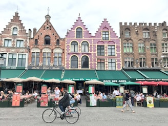 A vibrant European street scene featuring a row of colorful, historic buildings with intricate architectural details. The facades are decorated in shades of pink, purple, and earthy tones. Below the buildings, a bustling café area is filled with people sitting at tables under green awnings, enjoying food and drinks. In the foreground, a cyclist rides by, while other pedestrians walk along the cobblestone street.