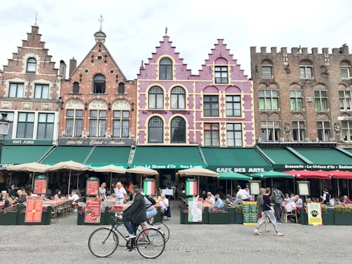 A vibrant European street scene featuring a row of colorful, historic buildings with intricate architectural details. The facades are decorated in shades of pink, purple, and earthy tones. Below the buildings, a bustling café area is filled with people sitting at tables under green awnings, enjoying food and drinks. In the foreground, a cyclist rides by, while other pedestrians walk along the cobblestone street.