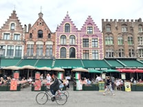 A vibrant European street scene featuring a row of colorful, historic buildings with intricate architectural details. The facades are decorated in shades of pink, purple, and earthy tones. Below the buildings, a bustling caf&eacute; area is filled with people sitting at tables under green awnings, enjoying food and drinks. In the foreground, a cyclist rides by, while other pedestrians walk along the cobblestone street.